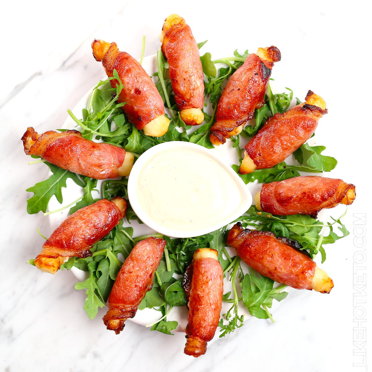 Bacon wrapped halloumi fried snacks on a large plate, surrounding a smalll bowl of ranch dipping sauce.