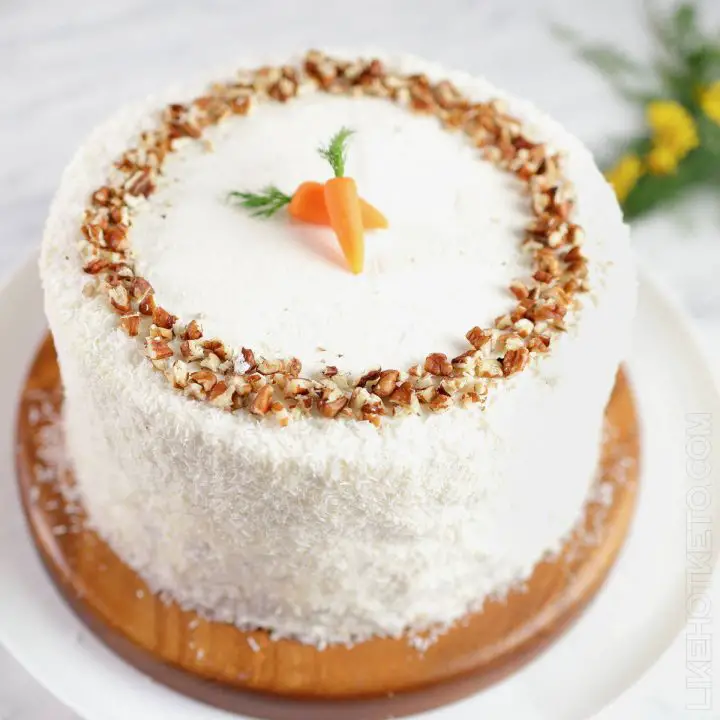 A tall, layered keto carrot cake frosted with cream cheese buttercream and decorated with grated coconut, baby carrots.
