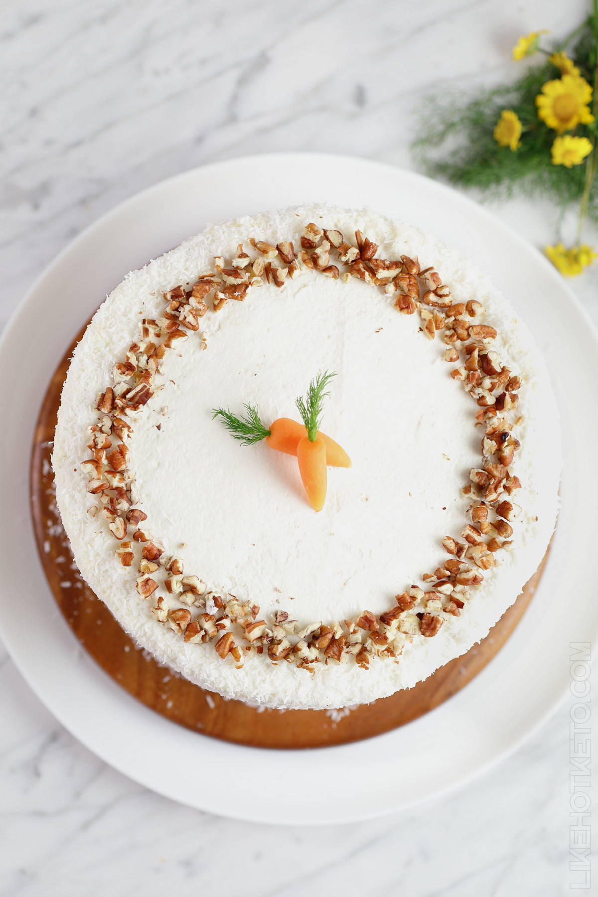 Keto carrot cake with white cream cheese frosting, fine shredded coconut and crushed pecans, with spring flowers on the side.