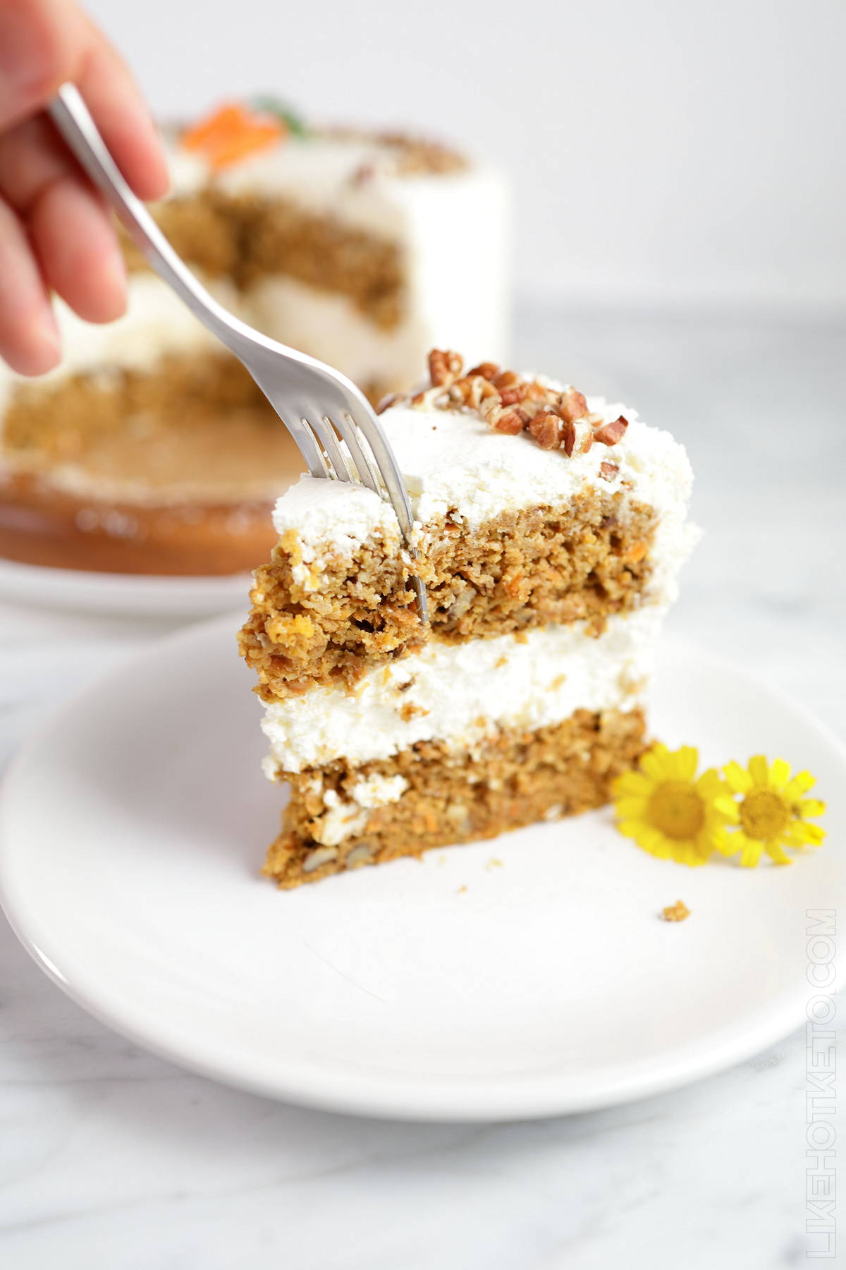 A fork cutting into a big slice of keto carrot cake with cream cheese frosting, on a white plate next to small yellow flowers, with the sliced cake on the background.