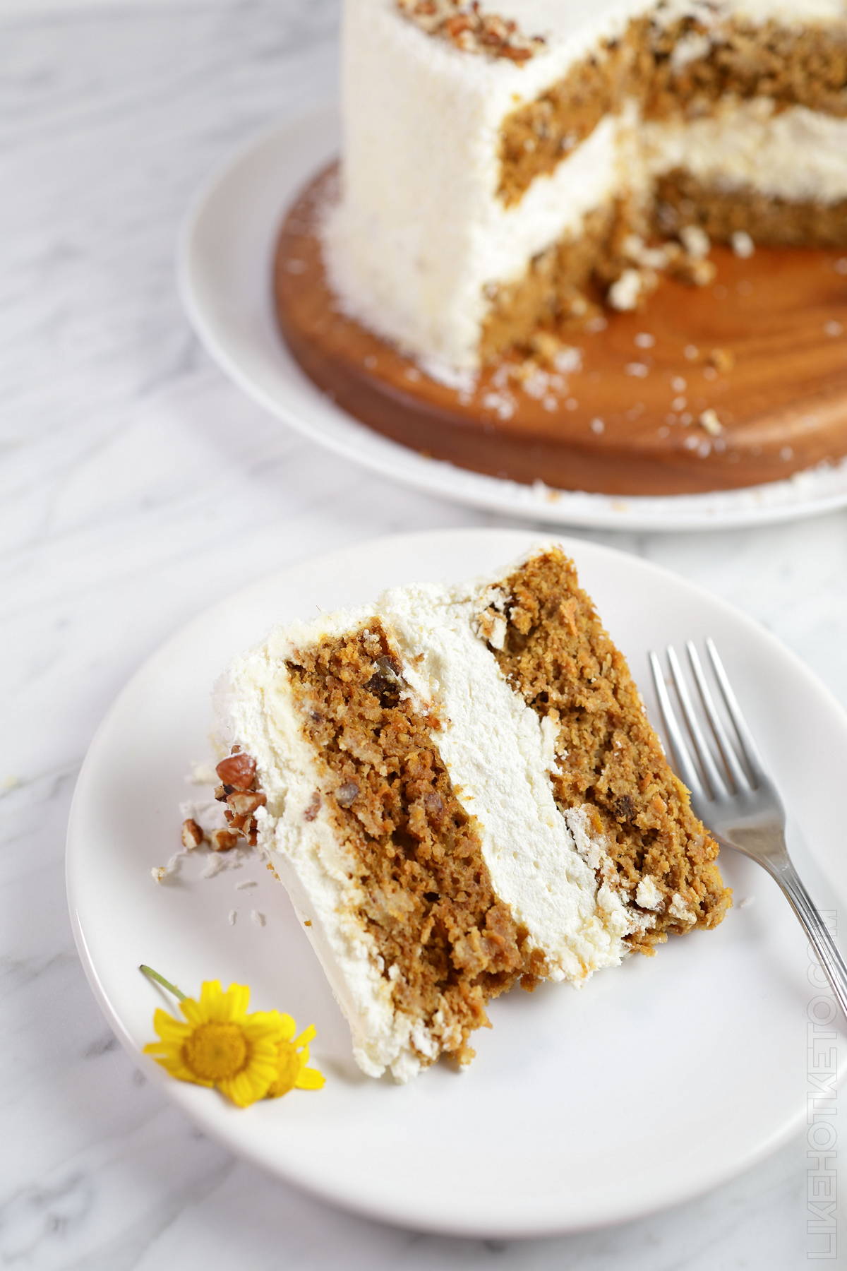 Slice of lupin flour carrot cake on a white plate, fork and flowers.