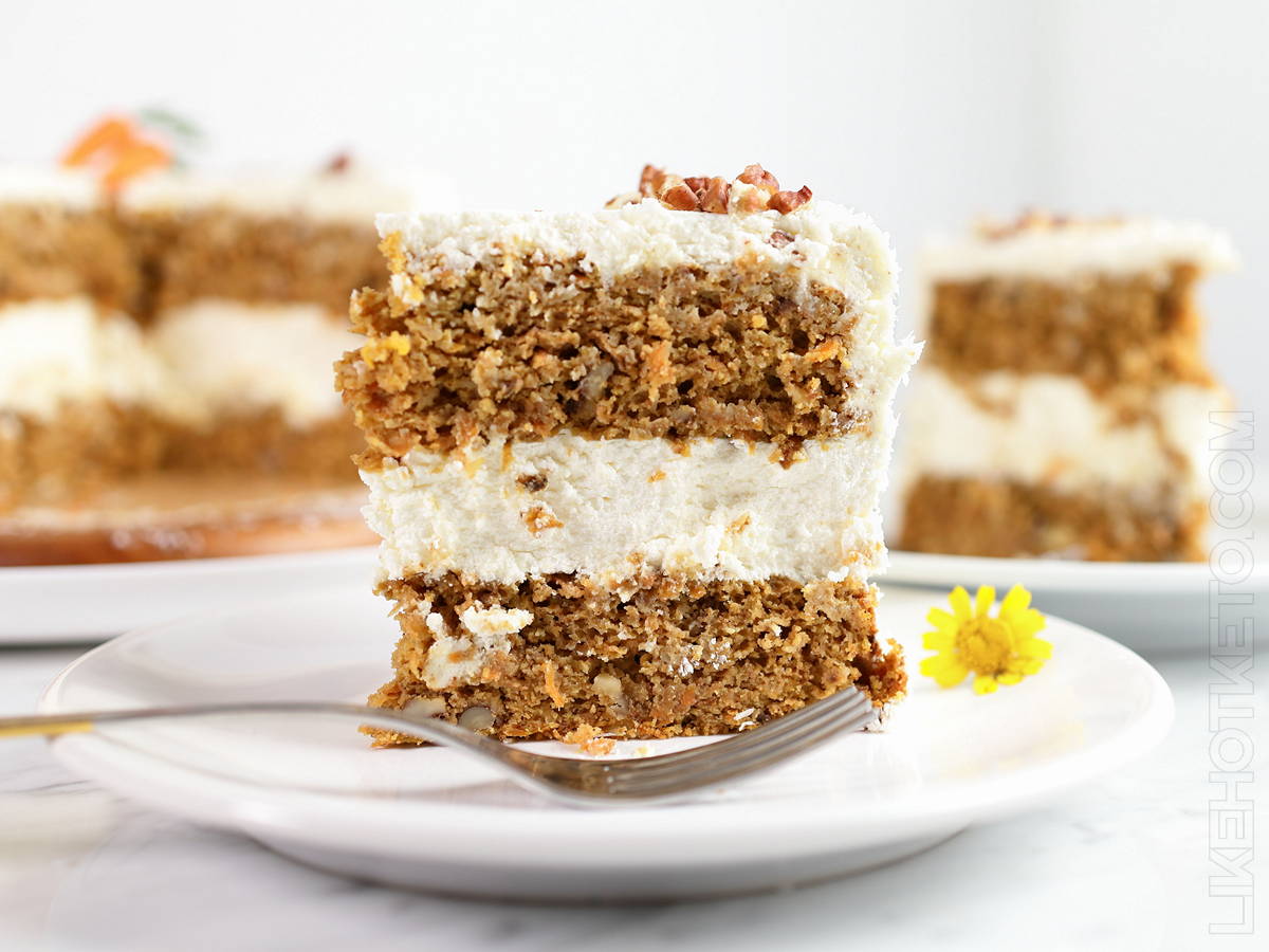 A slice of lupin flour carrot cake with coconut and pecans, on a white plate next to a silver fork.
