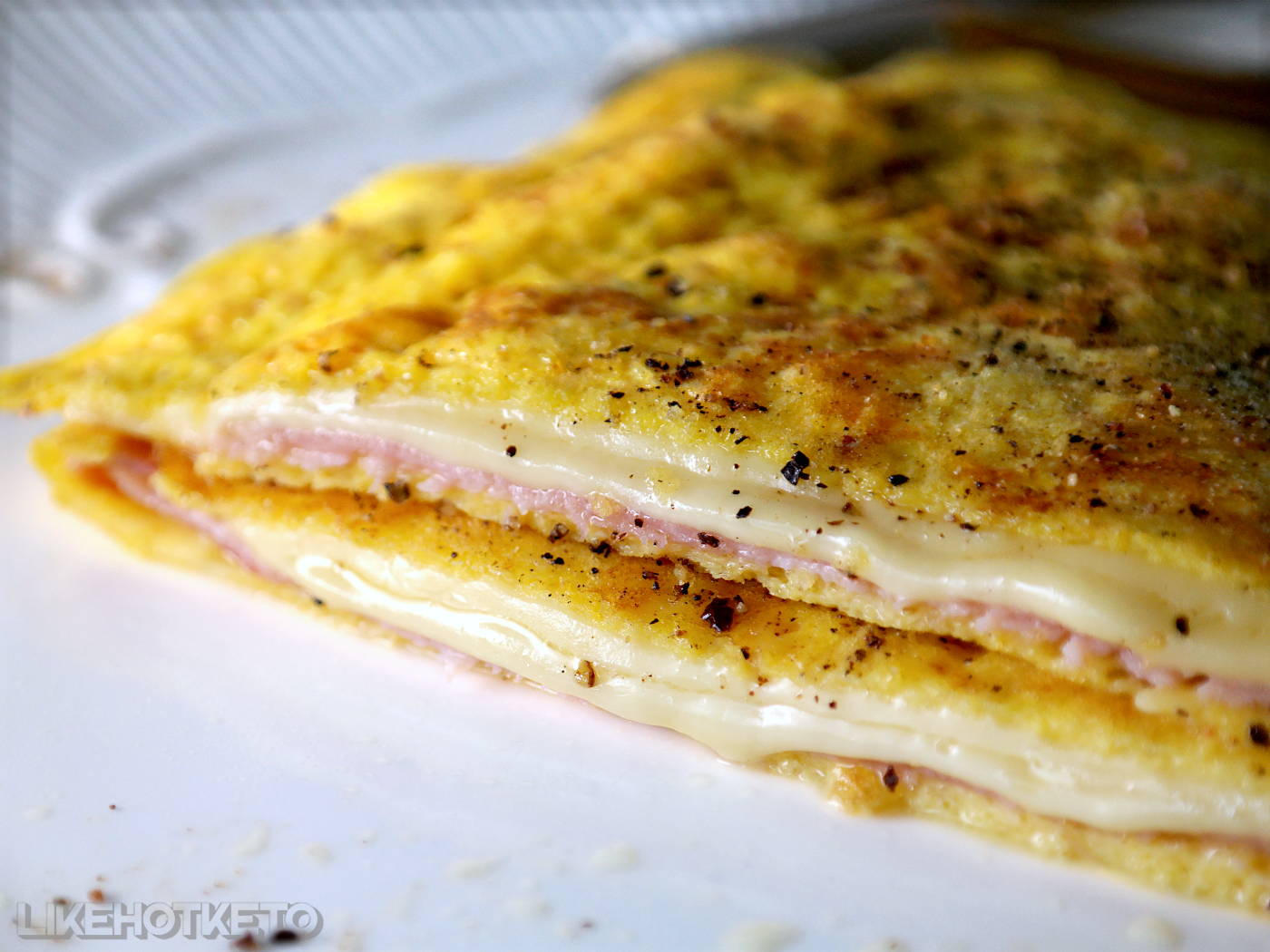 Keto egg crêpe folded on a white plate, cut in half, showing melted cheese and ham fillings.