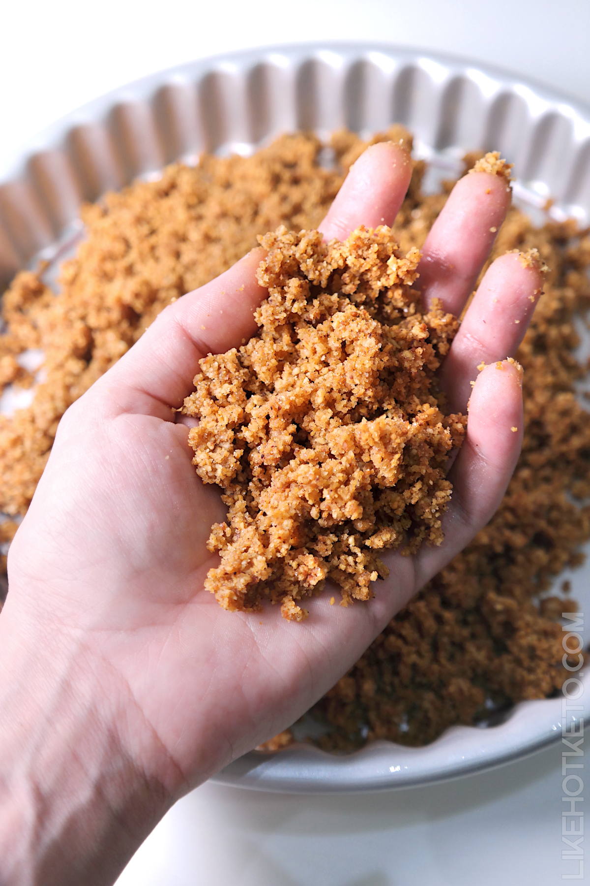 A hand full of keto Graham cracker moist crumbs, over a fluted pie pan with crust to be pressed.