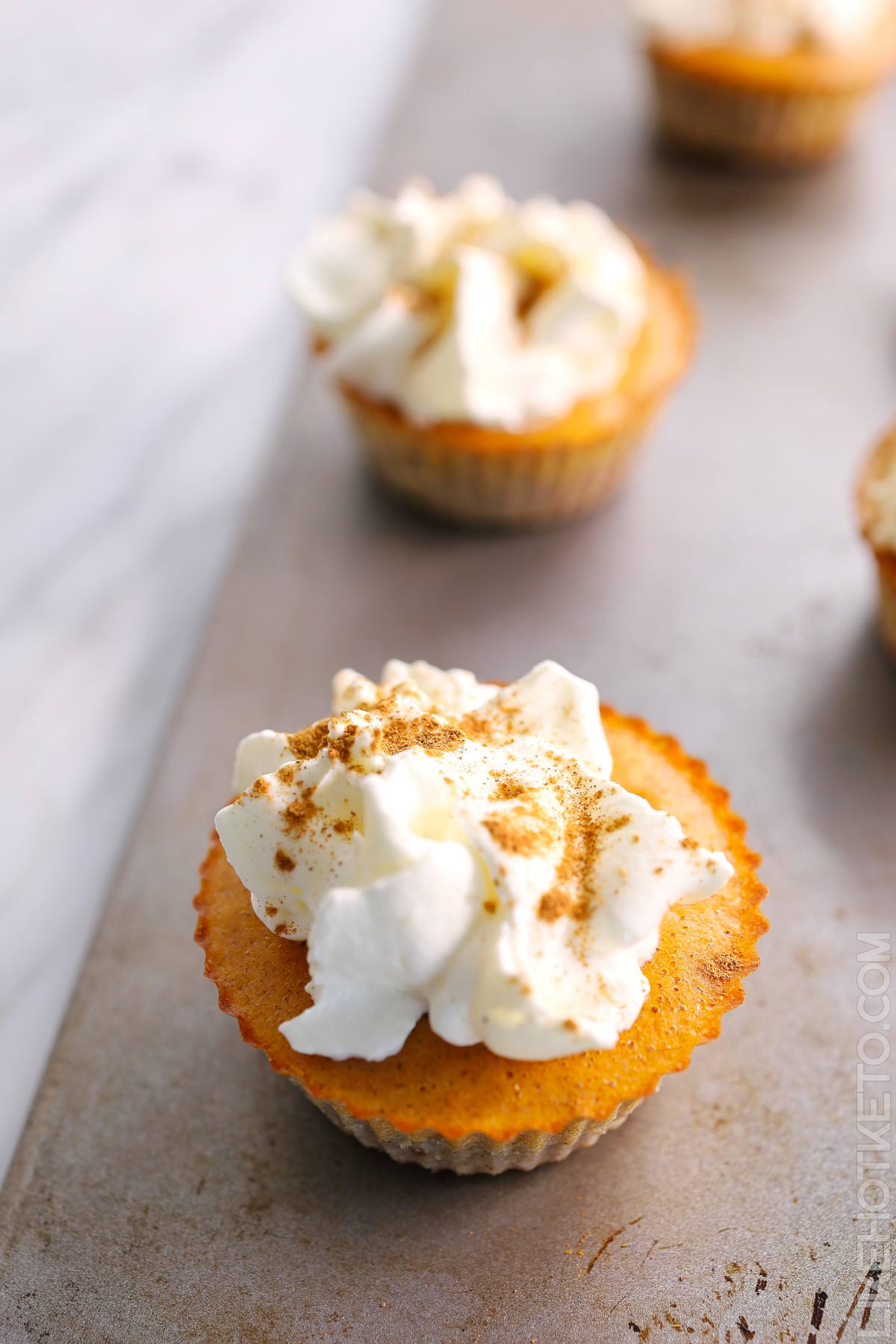 Baby keto pumpkin pies topped with whipped cream and cinnamon on a metal baking sheet.