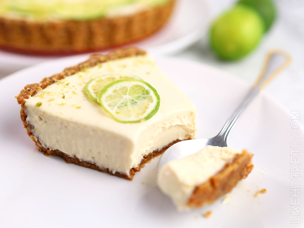 Slice of Key lime pie cheesecake on a white plate next to a couple of limes.