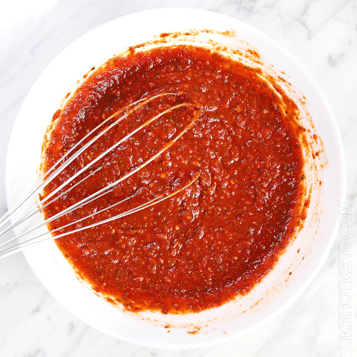 Sugar-free tomato sauce in white bowl with a large whisk.
