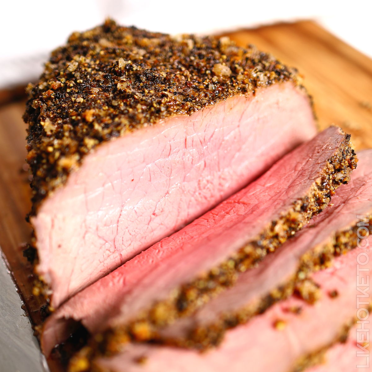 Peppercorn crusted beef roast, carved on wooden board, with medium-rare pink slices.