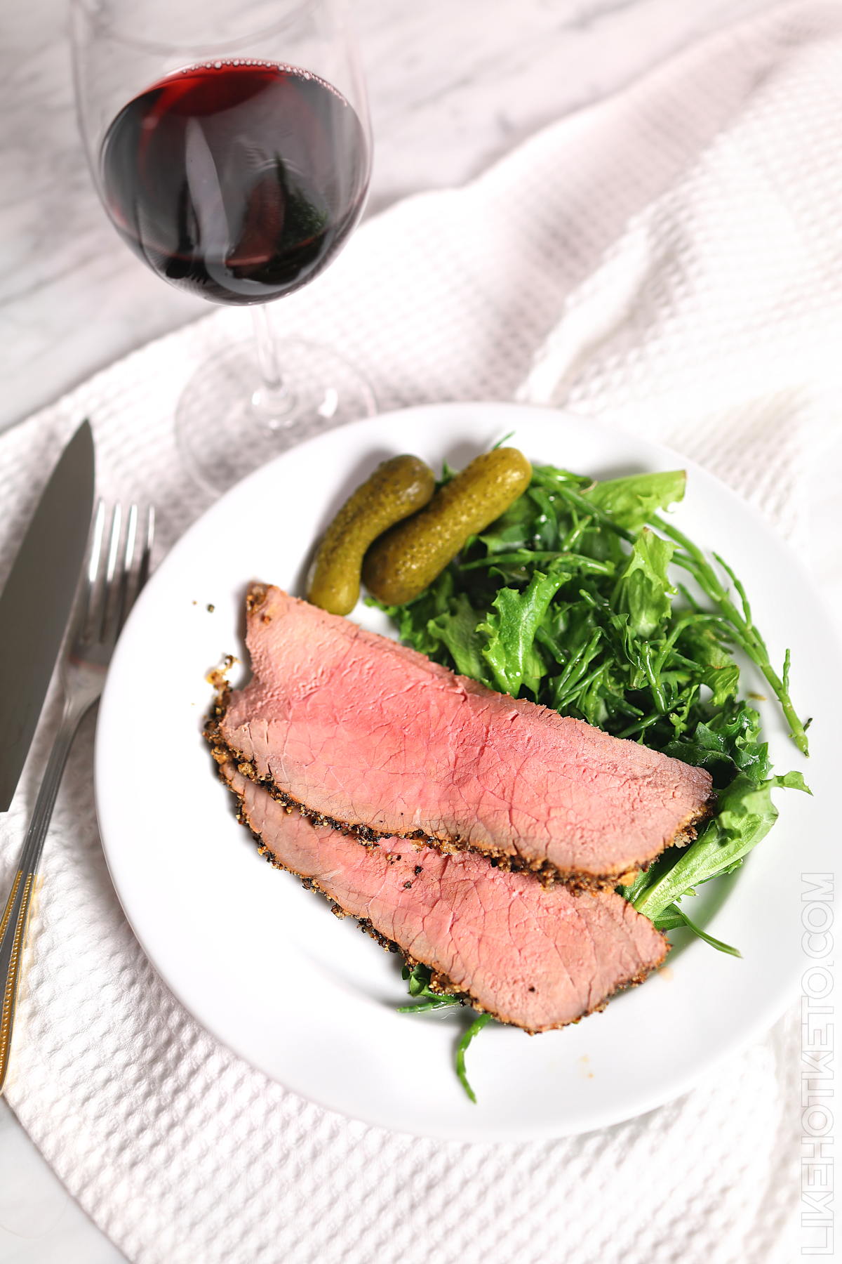 Keto roast beef dinner, with pickles and greens on a white plate, with a glass of red wine.