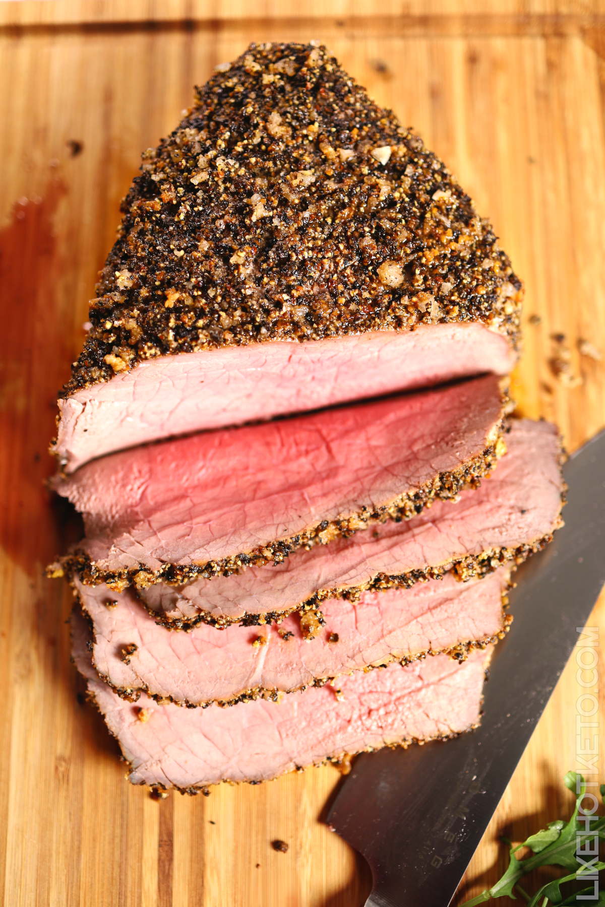 Half sliced roasted beef with thick crushed peppercorn crust,  on a wooden board next to cutting knife.
