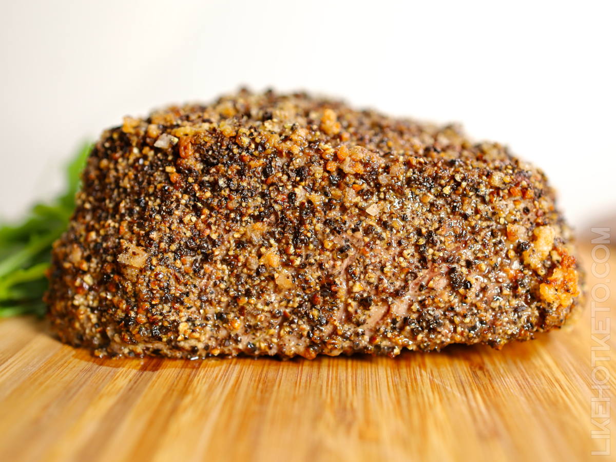 Side view of a peppercorn crusted beef roast resting on a wooden board.