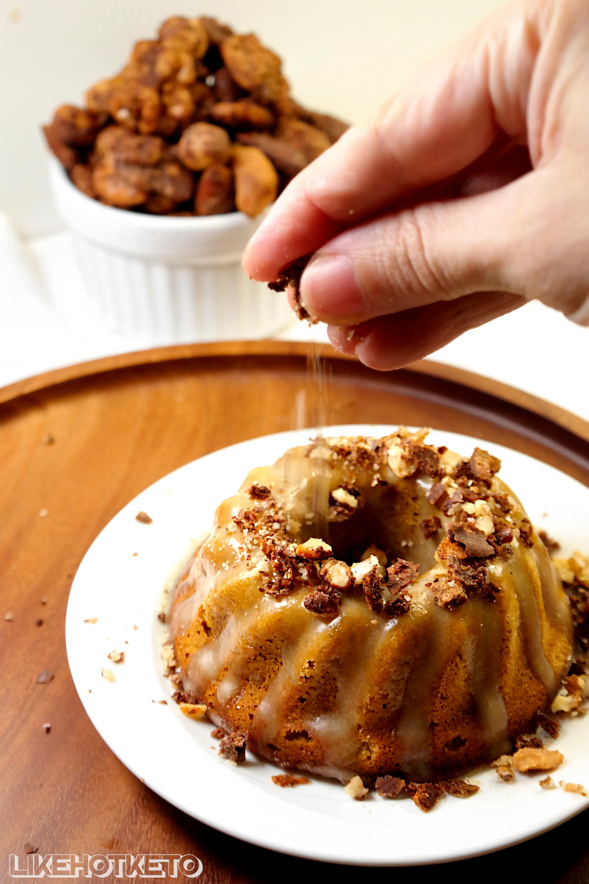 Crushed sugar-free candied nuts being sprinkled over a keto pumpkin pie spice mini bundt cake covered in cream cheese frosting.