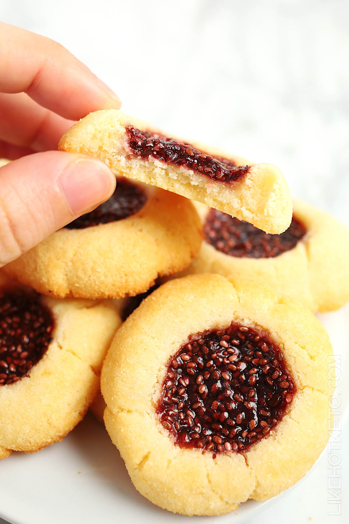 Half bitten berry chia jam thumbprint cookie, with buttery crumbs, over a plate with more thumbprint cookies piled up.
