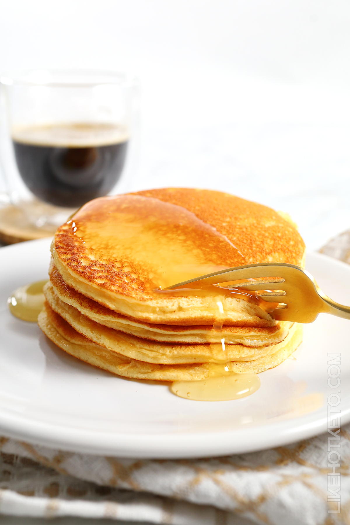 Stack of fluffy whey protein pancakes drizzled with sugar-free maple syrup being cut with a golden fork, a glass cup of espresso coffee in the background.