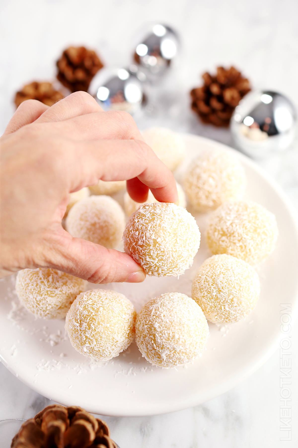 Hand reaching into a plate of snowball truffles surrounded with pine cones and Christmas balls.