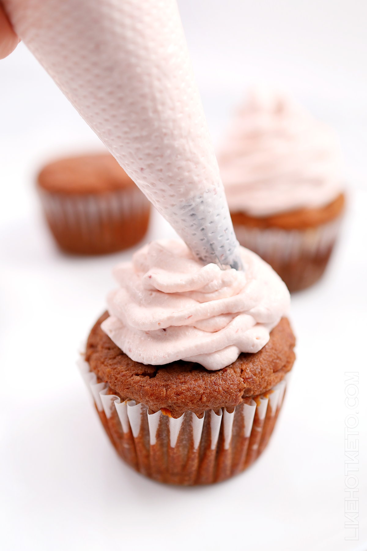 Applying  strawberry cream cheese frosting from a piping bag to a keto strawberry cupcake.