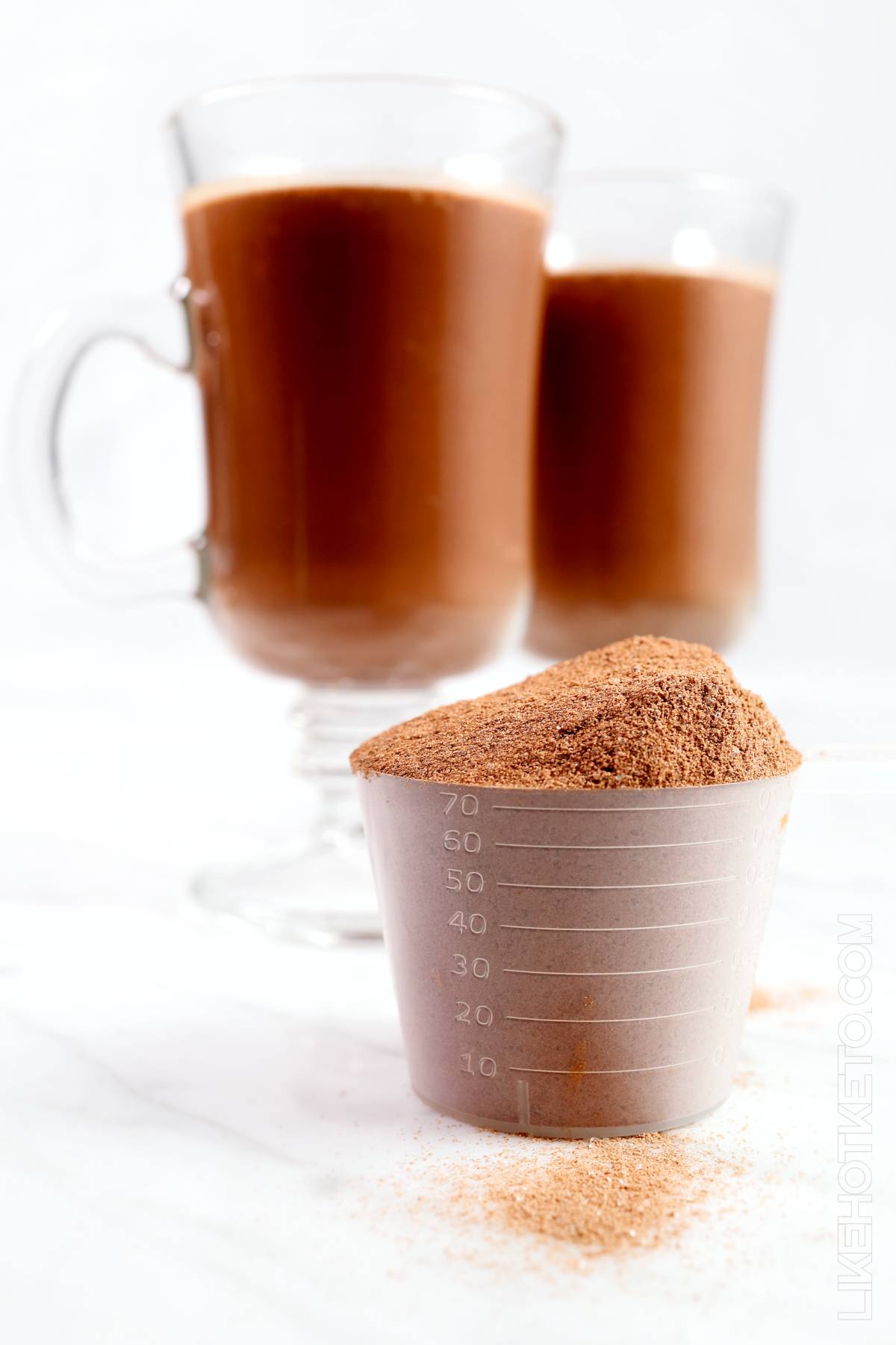 Protein measuring scoop filled with whey protein hot cocoa mix and two mugs of hot cocoa.